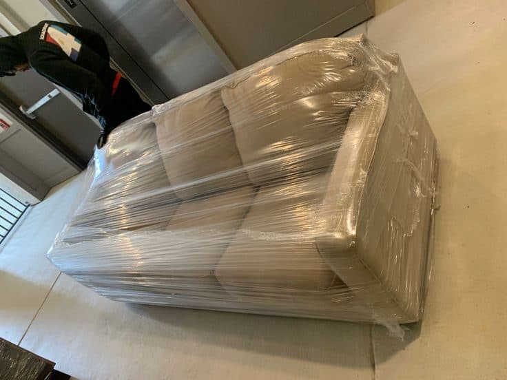 A large sofa fully wrapped in clear stretch film, moved through a building lobby on delivery day